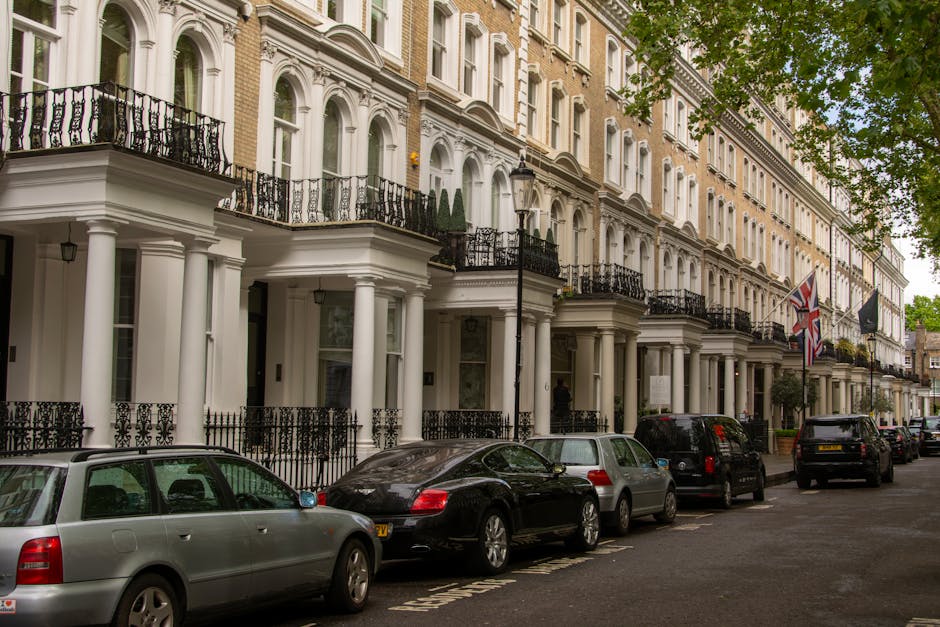 A row of elegant, multi-story Georgian-style terraced houses with cream-colored facades, black wrought iron balconies, and tall white columns supporting entrances, situated along a tree-lined street. 