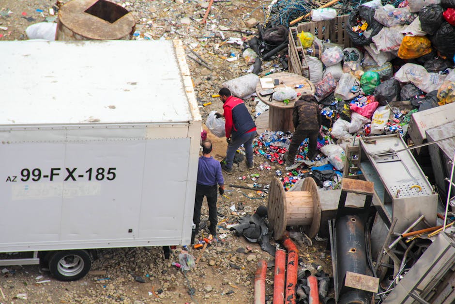 A large green waste disposal bin filled with a mixture of cardboard boxes, some flattened and others intact, along with a few smaller packaging items, is positioned on a city street. The bin appears w