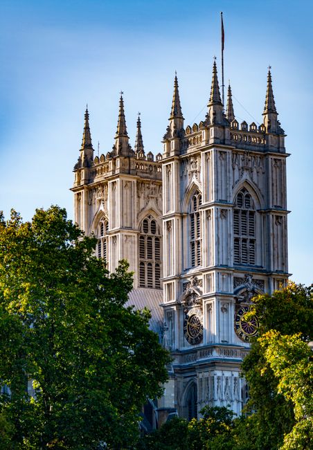 A tall, historic stone clock tower with multiple narrow, arched windows and intricate architectural details, standing prominently against a bright blue sky. The tower features four smaller spires at e