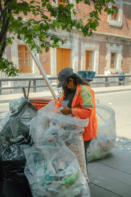 A large green wheelie bin positioned on a paved surface beside a dark brick wall. The bin is filled with various red and black bags of construction or garden waste, some labeled with specifications su